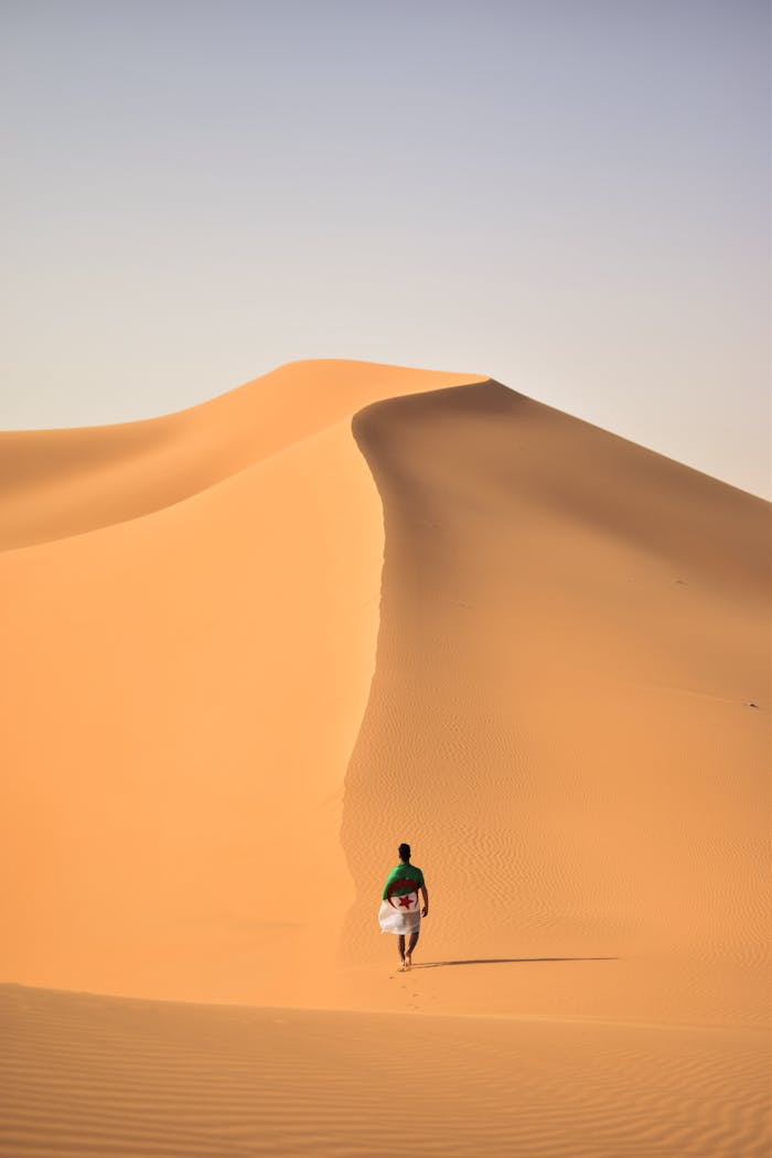 A lone traveler walking over the vast, golden sand dunes of the Sahara Desert in Algeria.