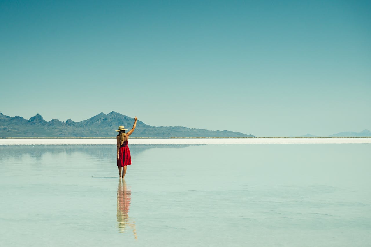 A woman in a red dress stands at a serene salt flat with mountains in the background, enjoying solitude and reflection.